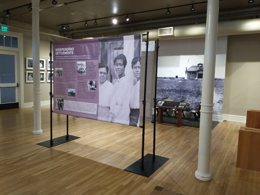 Free-standing banner stands with museum text and black and white photo in a room with wooden floors.