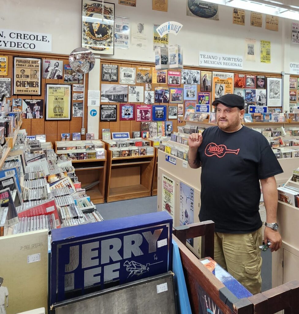 A man with a black tshirt and black cap stands inside of a record store.