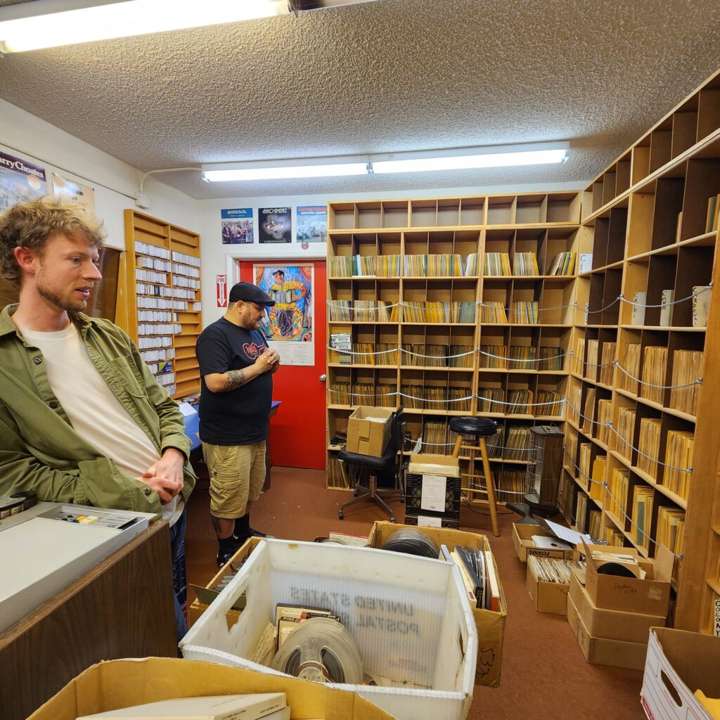 Two men stand inside a record archive with records organized on shelves