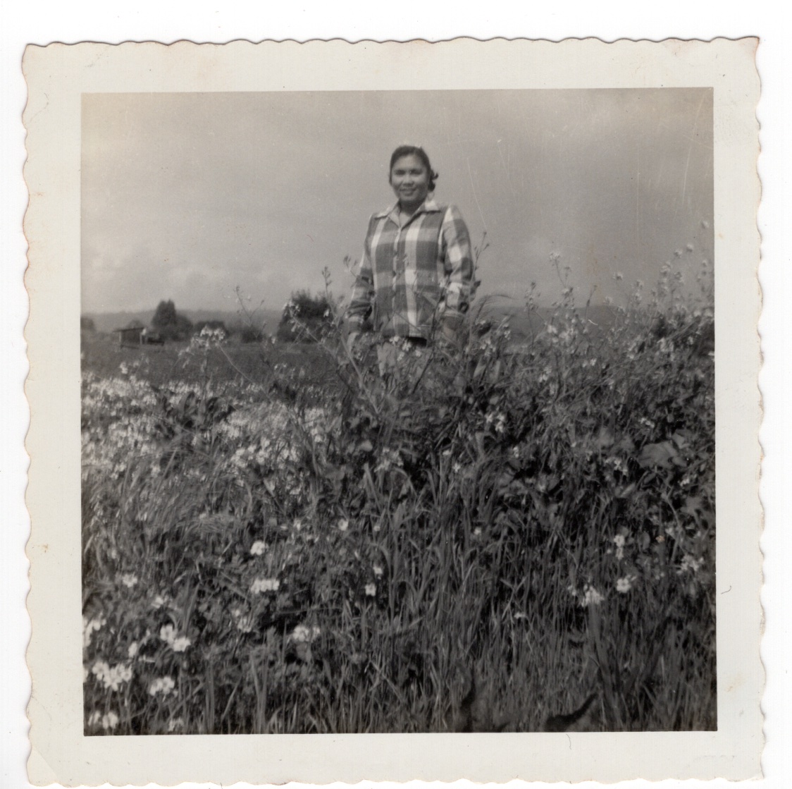 Square black and white photograph of a woman wearing a plaid shirt, standing in a field of wildflowers.