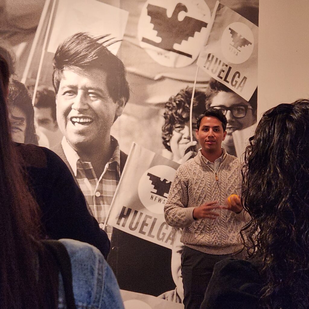 Man wearing white textured sweater and grey slacks stands in front of a large photo mural of Cesar Chavez