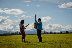Two people wearing light blue denim button shirts holding guitars stand in a field of grass, mountains and clouds in the background.