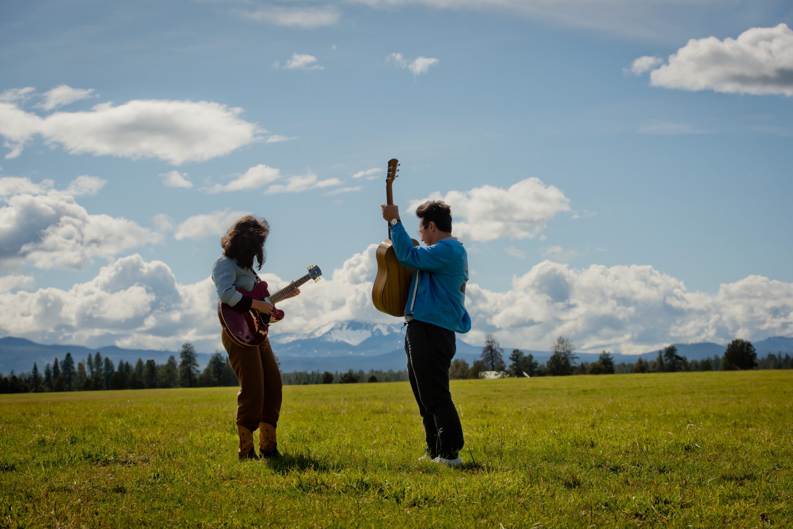 Two people wearing light blue denim button shirts holding guitars stand in a field of grass, mountains and clouds in the background.