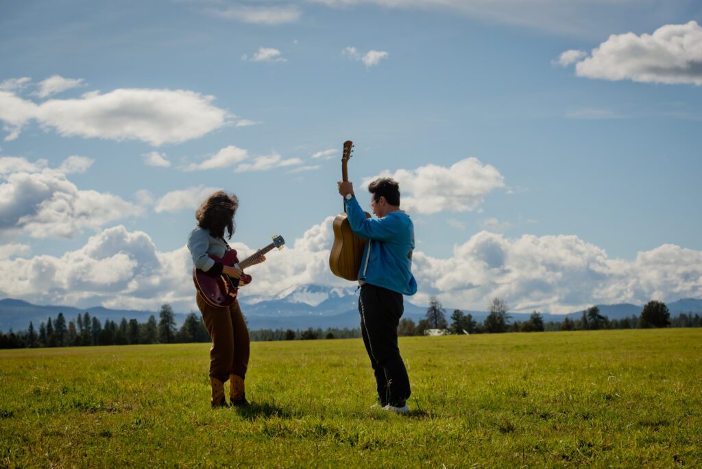 man and woman wearing light blue longsleeve shirts hold acoustic and electric guitars in an open field, with puffy clouds in the distance.