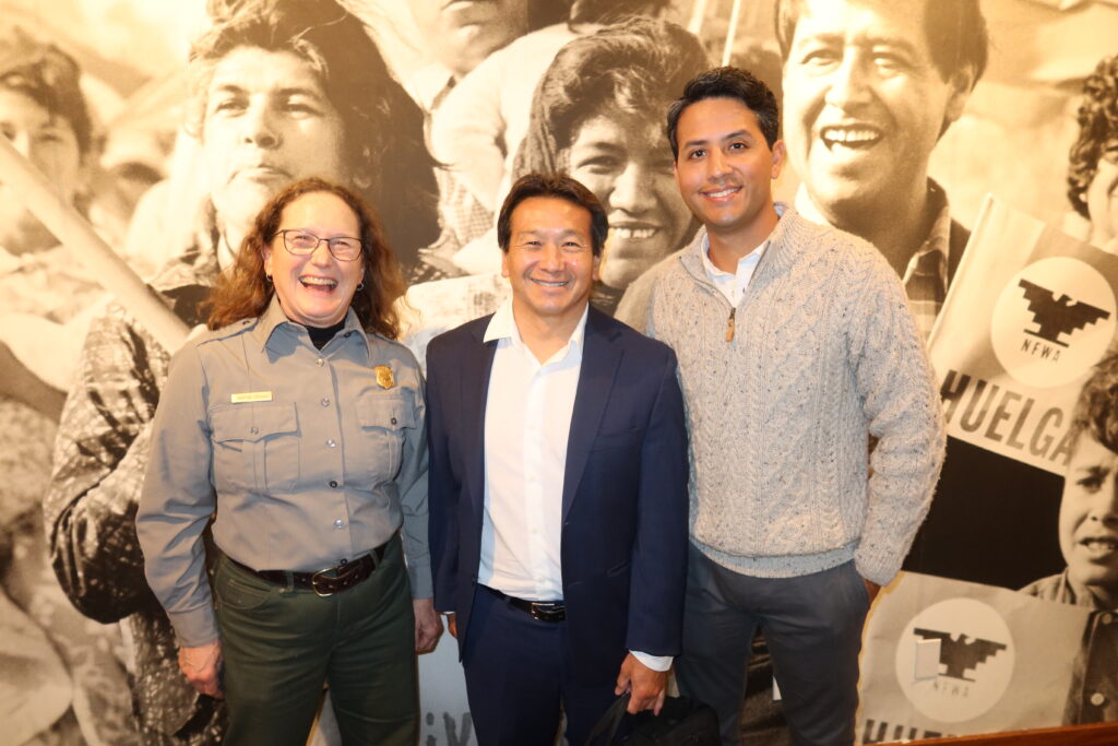 Woman wearing National Park Service uniform poses for a photo with a man wearing white dress shirt and dark blue suit jacket and man wearing beige cable knit sweater