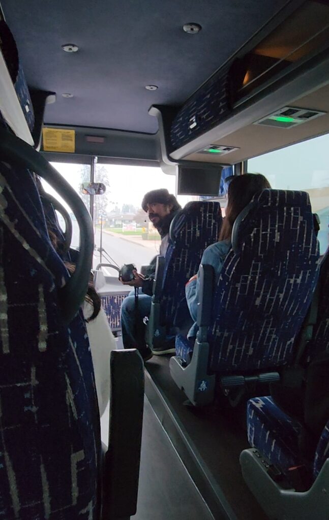 Man wearing portable speaker sits at front of a large tour bus and looks back at seated passengers while speaking