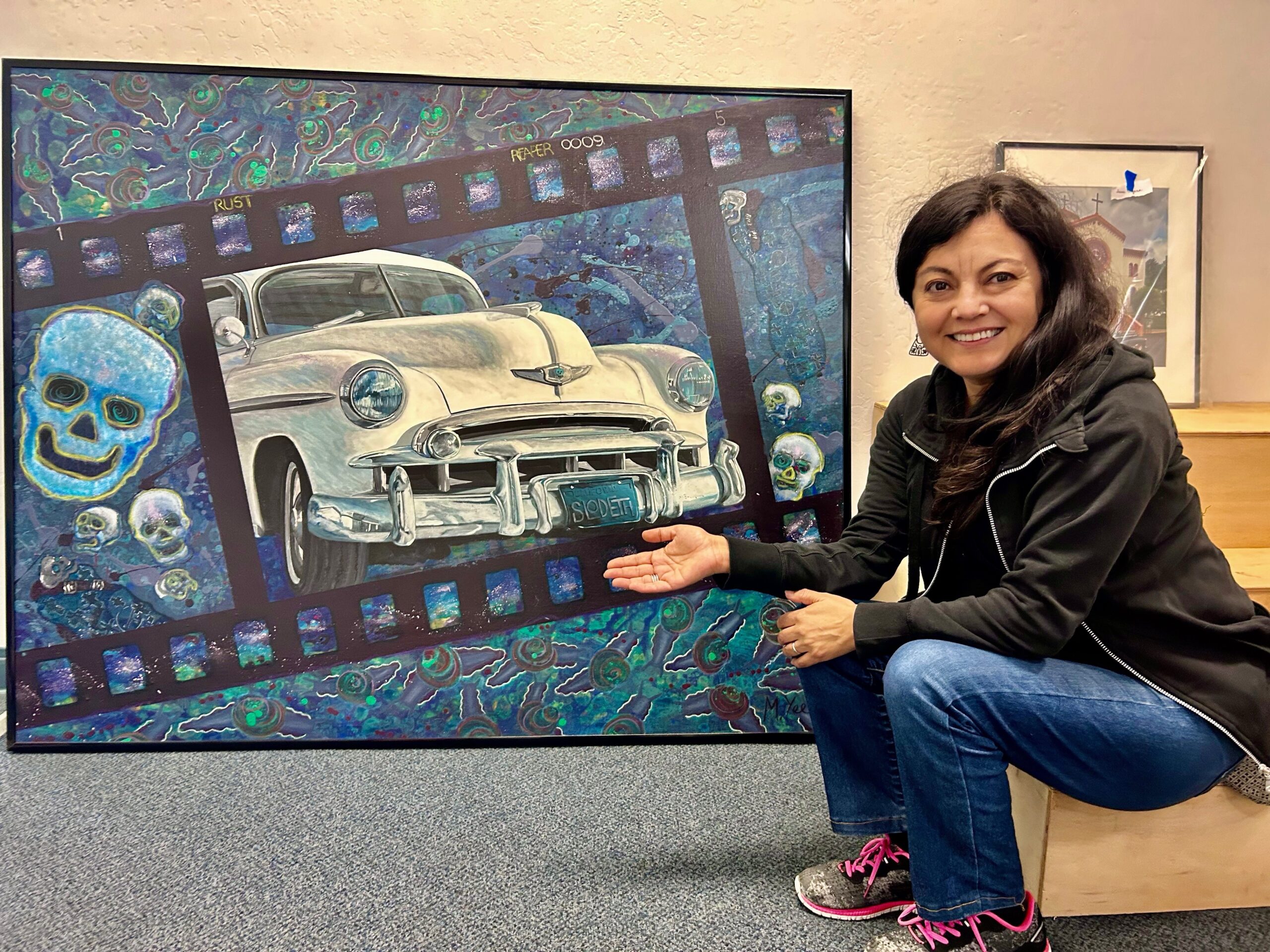 Woman wearing black jacket and jeans crouching for a photo next to piece of artwork featuring a lowrider card.