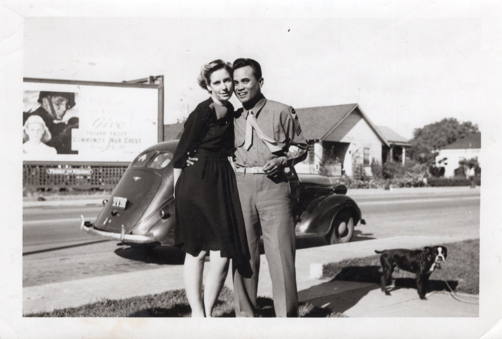 Black and white photo of a couple in the 1940s, standing in front of a car and billboard