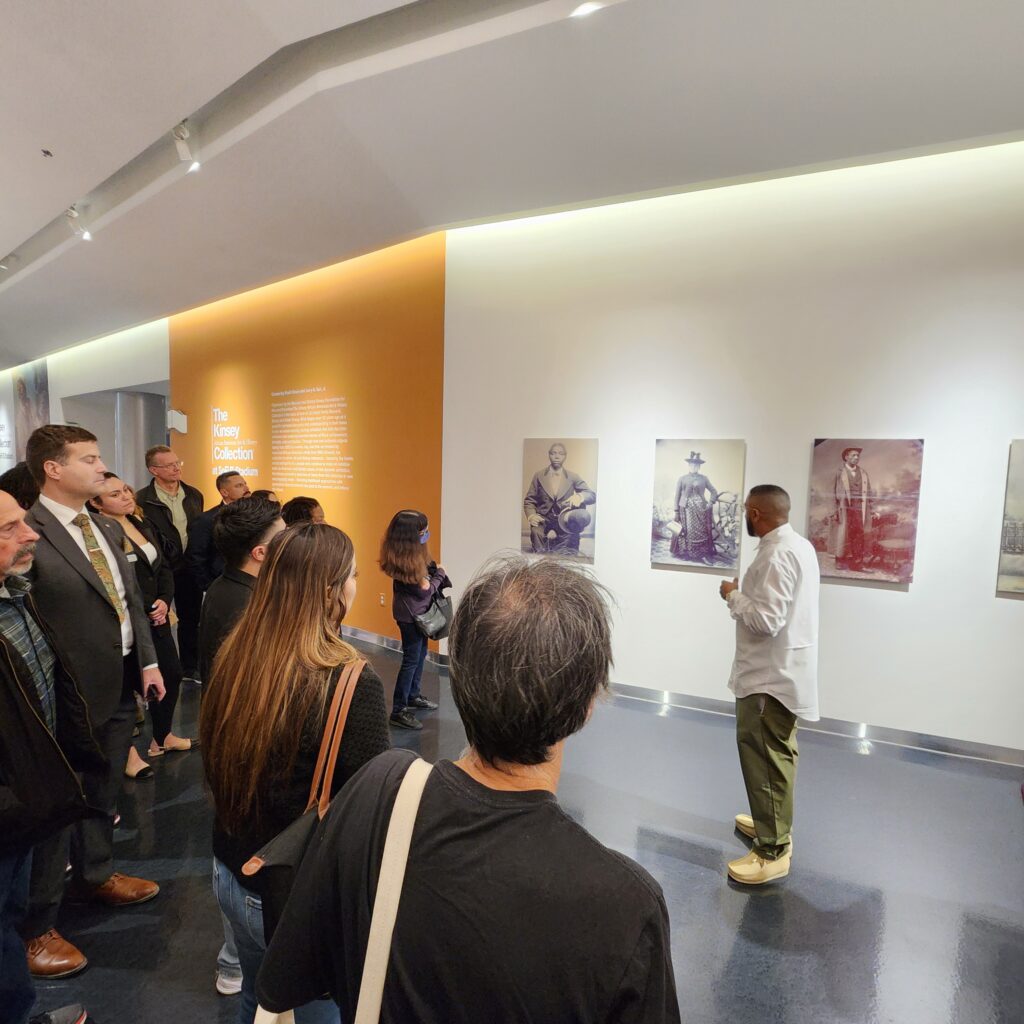 Man wearing longsleeve white shirt and khaki pants speaks to a crowd inside a gallery