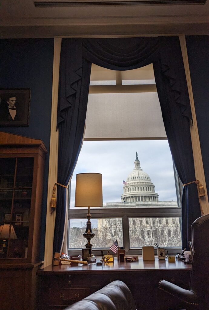 View from inside an office with leather couch and desk lamp in the foreground, with view of US Capitol dome outside the window.