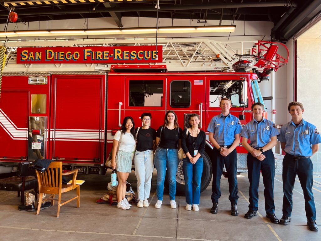 Group of four students pose for photo with three fire and rescue staff in uniform, in front of a red fire truck/engine