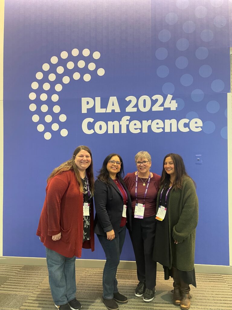 group of four women pose for a photo in front of a wall reading "PLA 2024 Conference