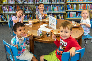 Group of five children sit across a low table with lunches, inside a library.