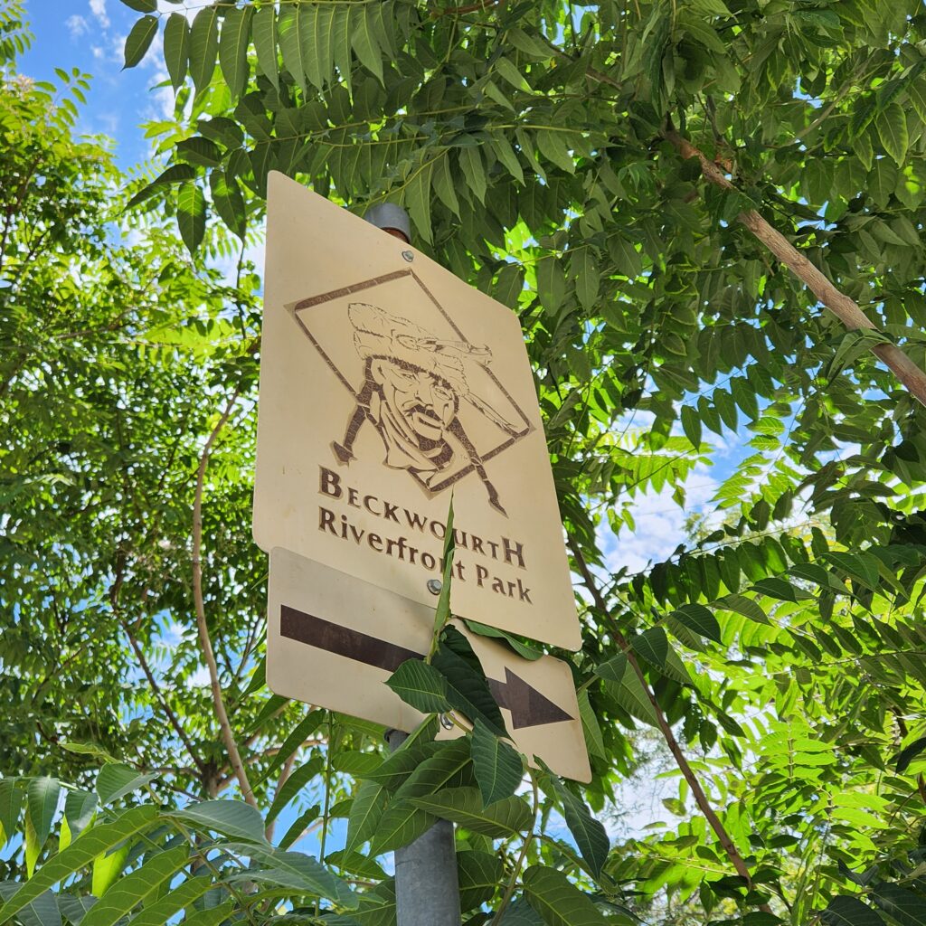 Place marker sign in tan outside amidst trees. Text reads James Beckfourth Riverfront Park.