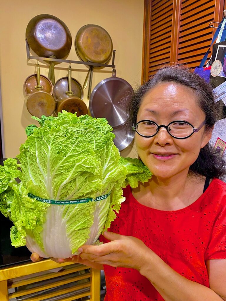 Photo of a woman wearing a red top and black rimmed glasses holding up a large cabbage.