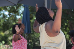 Two women participate in a wellness exercise outside, with arms raised towards the sky.