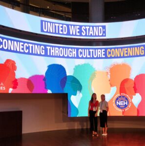 Two women pose for a photo in front of a screen reading "United We Stand: Connecting Through Culture"