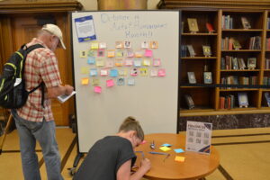 Man wearing flannel shirt and backpack facing a white board with post-it notes on it, title written in marker at the top reading October is Arts and Humanities Month