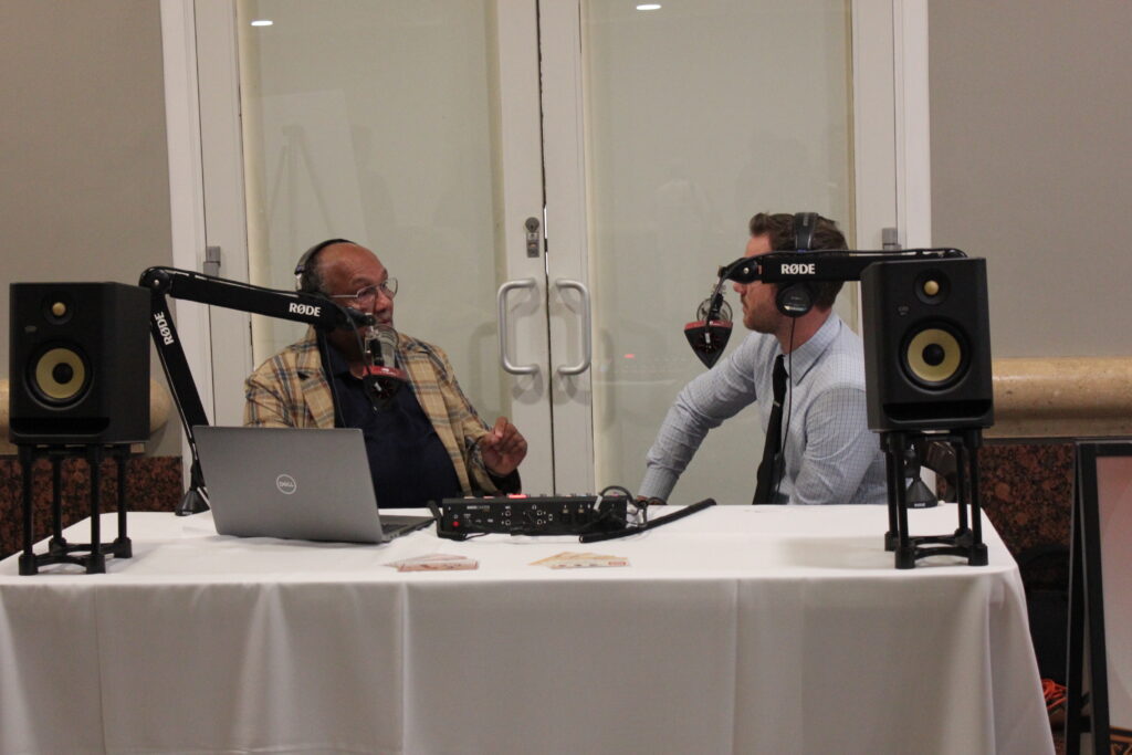 Two people sit behind podcasting equipment for an audio interview