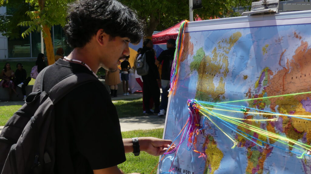 Student wearing black tshirt an black backpack looks a map of the world with pins and strings of yarn pinned across it.